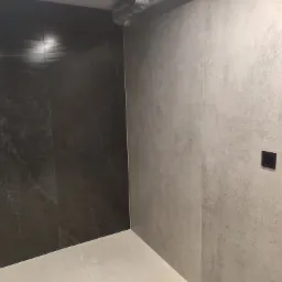 Wnętrze łazienki w trakcie prac wykończeniowych, widoczne ciemne i jasne płytki na ścianach, szara podłoga oraz czarny włącznik światła na jasnej ścianie.