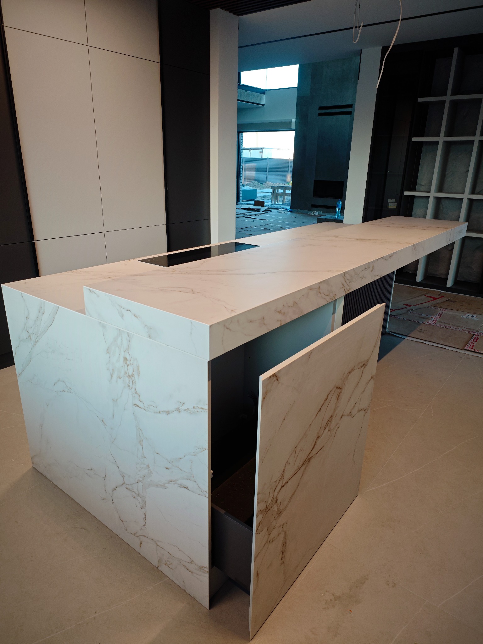 Wyspa kuchenna z blatem ze spieku kwarcowego o marmurkowym wzorze, z otwartą szafką i widocznym fragmentem płyty indukcyjnej. W tle okno i elementy wykończenia.