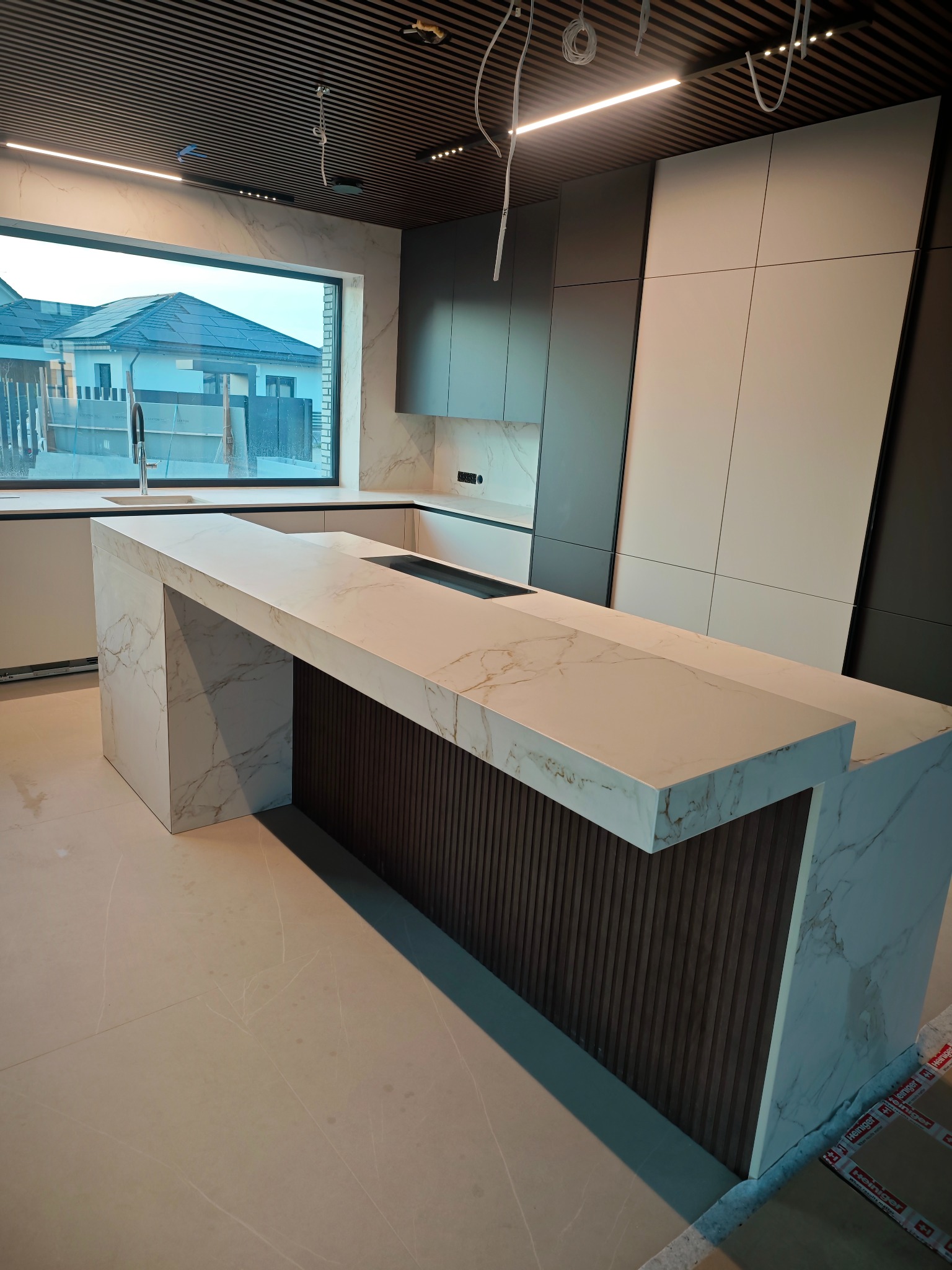 Nowoczesna kuchnia z wyspą i blatem ze spieku kwarcowego w odcieniach bieli i szarości. Minimalistyczny design, eleganckie wykończenie, widok na dom zza okna.