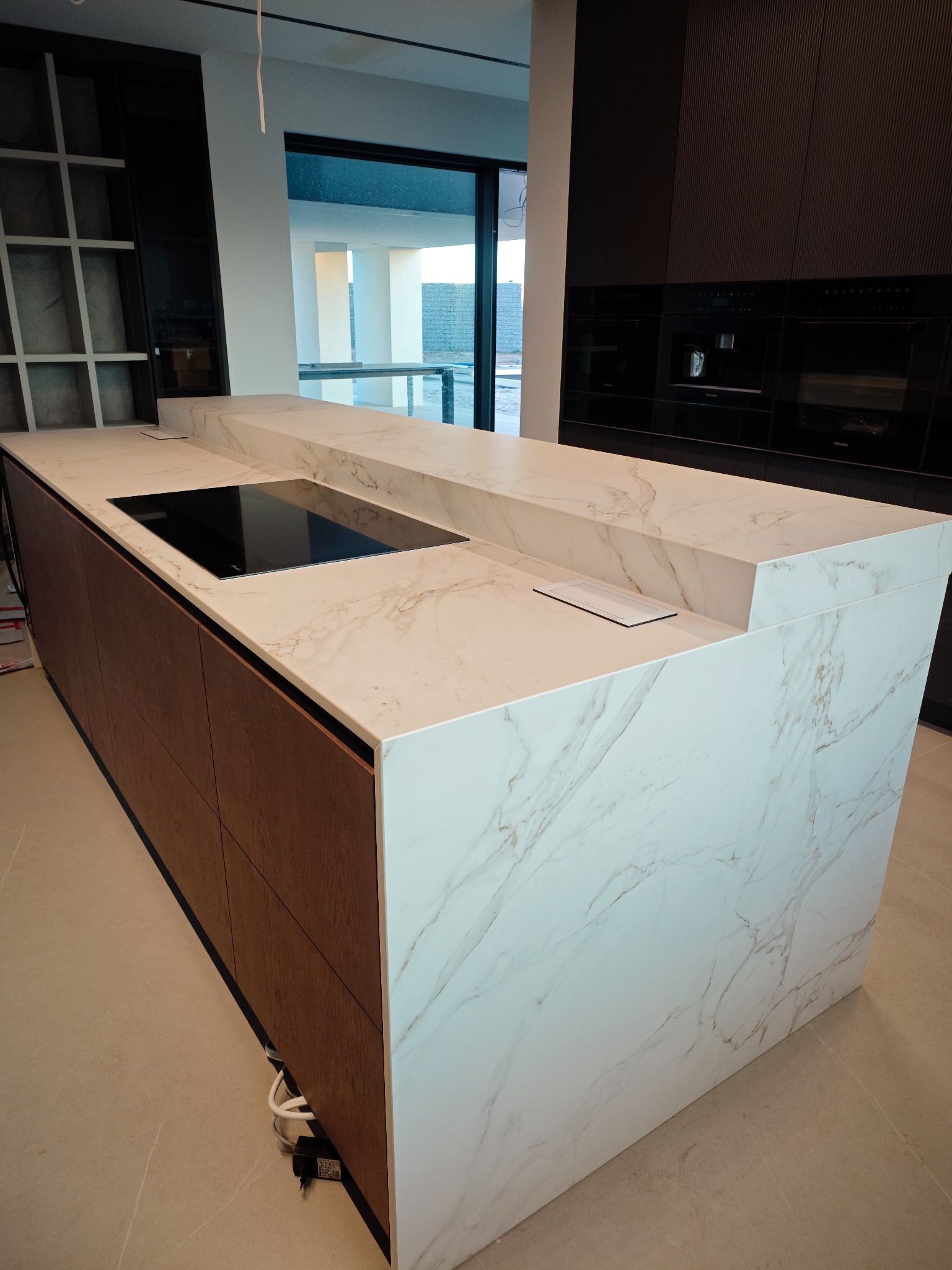 Wyspa kuchenna z blatem ze spieku kwarcowego o marmurkowym wzorze, z wbudowaną płytą indukcyjną i gniazdkami. Nowoczesne wykończenie wnętrza kuchni.