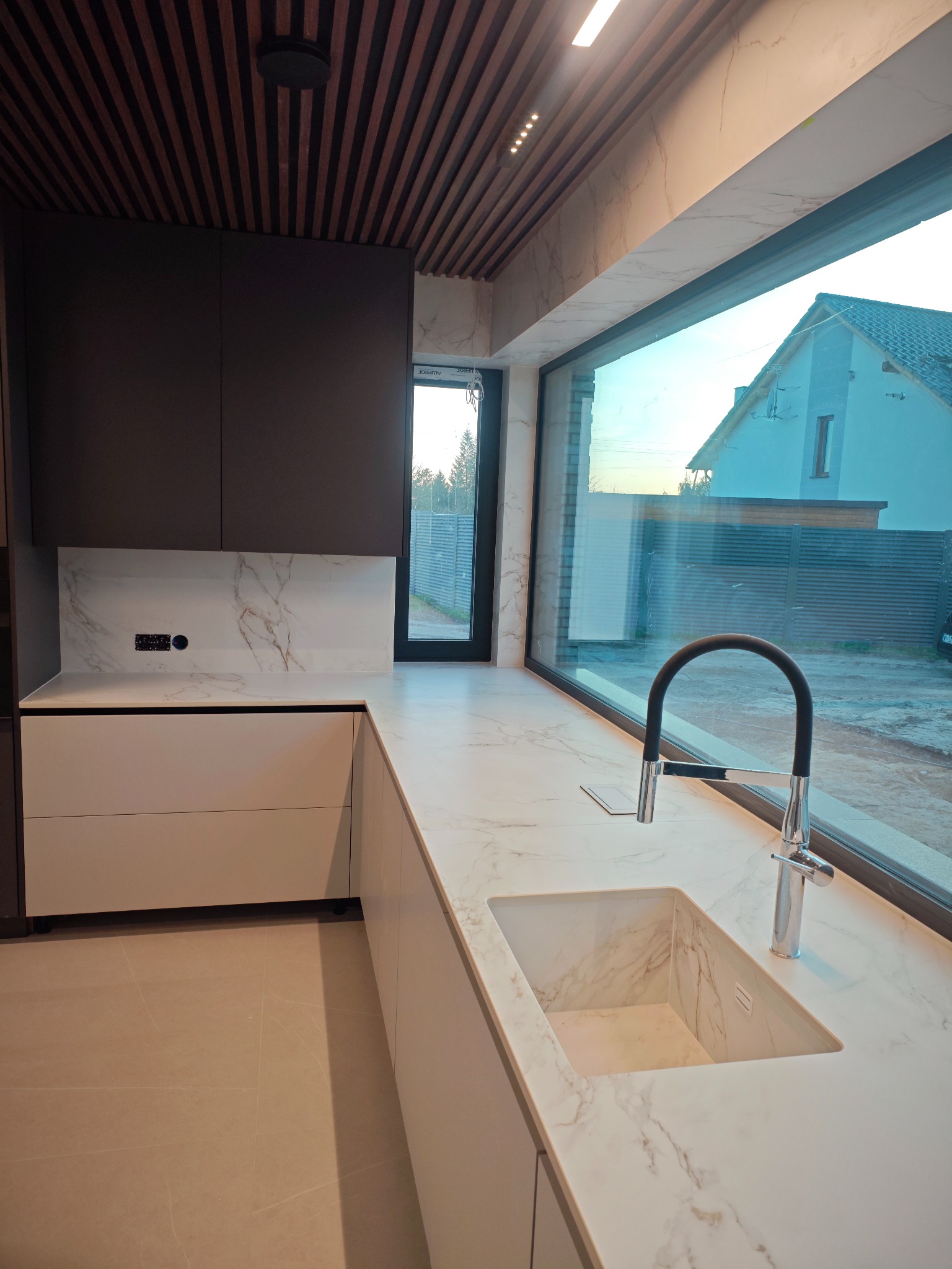 Nowoczesna kuchnia z blatem i zlewem ze spieku kwarcowego, z dużym oknem i drewnianym sufitem lamelowym. Minimalistyczny design, marmurowy wzór na blacie.