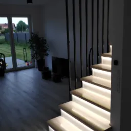 Nowoczesne schody drewniane z podświetleniem LED, czarne barierki pionowe, widok z dołu na jasne wnętrze domu z oknem na ogród.