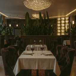Elegancka restauracja: stół z nakryciem, wino w kieliszkach, pluszowe krzesła, rośliny doniczkowe i dekoracyjne żyrandole. Ciepłe, stonowane oświetlenie.