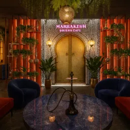 Wnętrze shisha cafe w stylu marokańskim: złote drzwi z ornamentami, rośliny, stoliki z fajkami wodnymi, miękkie sofy i fotele w ciepłej kolorystyce.