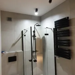 Nowoczesna łazienka z kabiną prysznicową typu walk-in z czarnymi profilami, wiszącą toaletą i grzejnikiem drabinkowym w kolorze czarnym, oświetlenie punktowe na suficie, szare płytki.