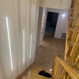 Sebastian Matysiak - Klatka schodowa z drewnianymi schodami i parkietem ułożonym w jodełkę. Na ścianie pionowe, wpuszczone oświetlenie LED. Widok z góry na dół.