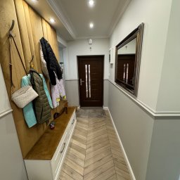 Sebastian Matysiak - Nowoczesny przedpokój z panelami ściennymi, lustrem i podłogą w jodełkę. Wieszaki z ubraniami i torebką, siedzisko z szufladami. Oświetlenie sufitowe.