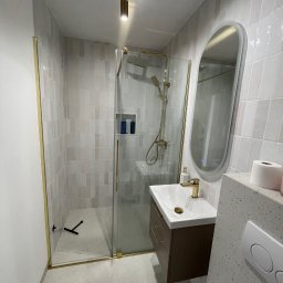 Sebastian Matysiak - Wykończona łazienka z kabiną prysznicową typu walk-in, złotymi detalami i owalnym lustrem. Białe płytki na ścianach i podłoga w jasnym lastryko.