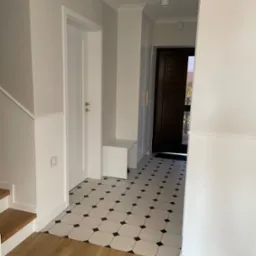 Jasne wnętrze holu z białą stolarką, panelami ściennymi i podłogą w biało-czarną szachownicę. Widoczne schody z drewnianymi stopniami i ciemne drzwi wejściowe.