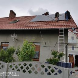 Solar Tech - Montaż paneli słonecznych na dachu domu z czerwonej dachówki, widoczni trzej monterzy przy pracy, drabina oparta o ścianę, na pierwszym planie betonowy płot z roślinnością.
