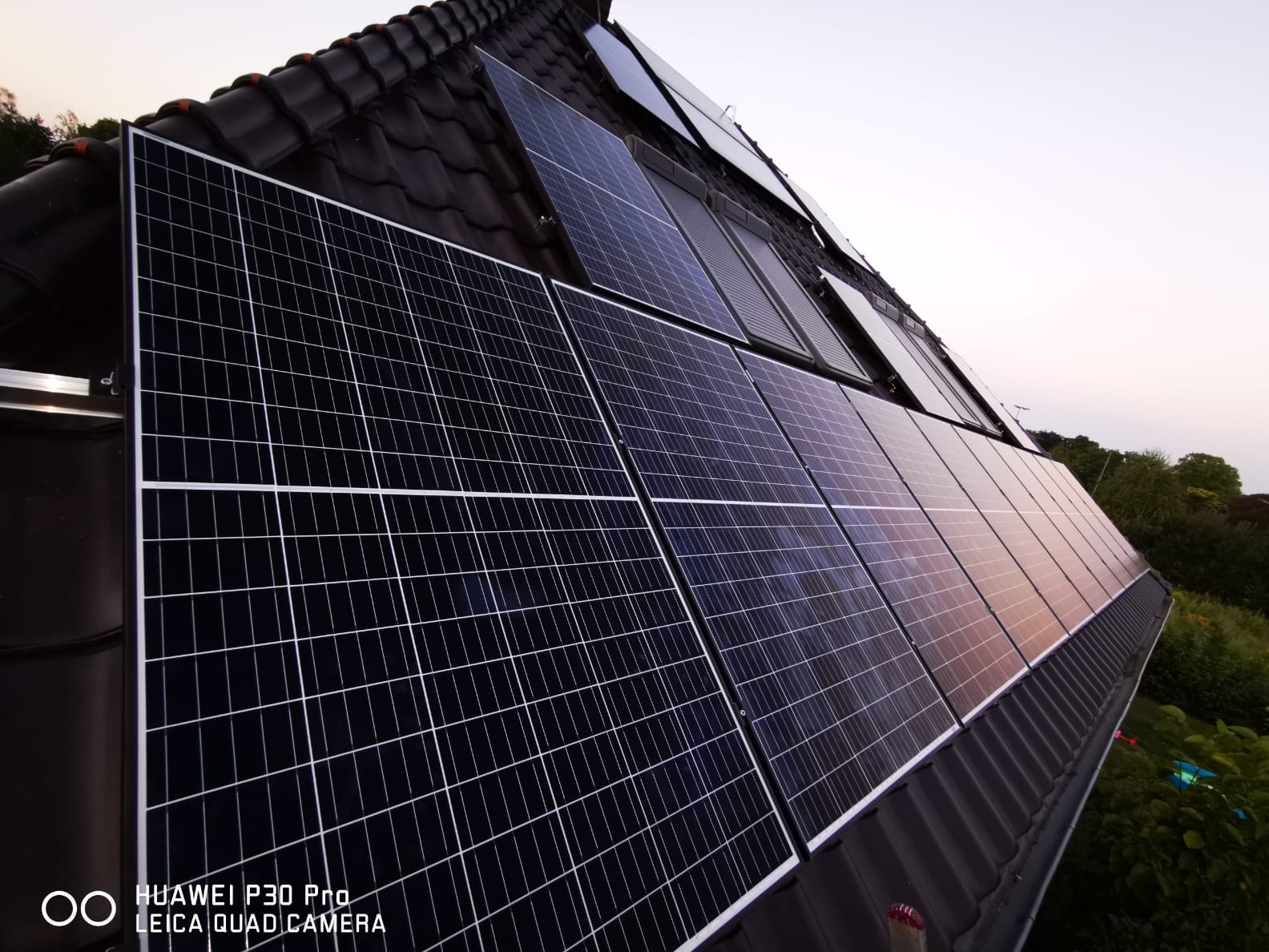 Panele fotowoltaiczne zamontowane na ciemnym, spadzistym dachu pokrytym dachówką, widoczne z dołu pod kątem, z zielenią drzew w tle i wieczornym niebem.