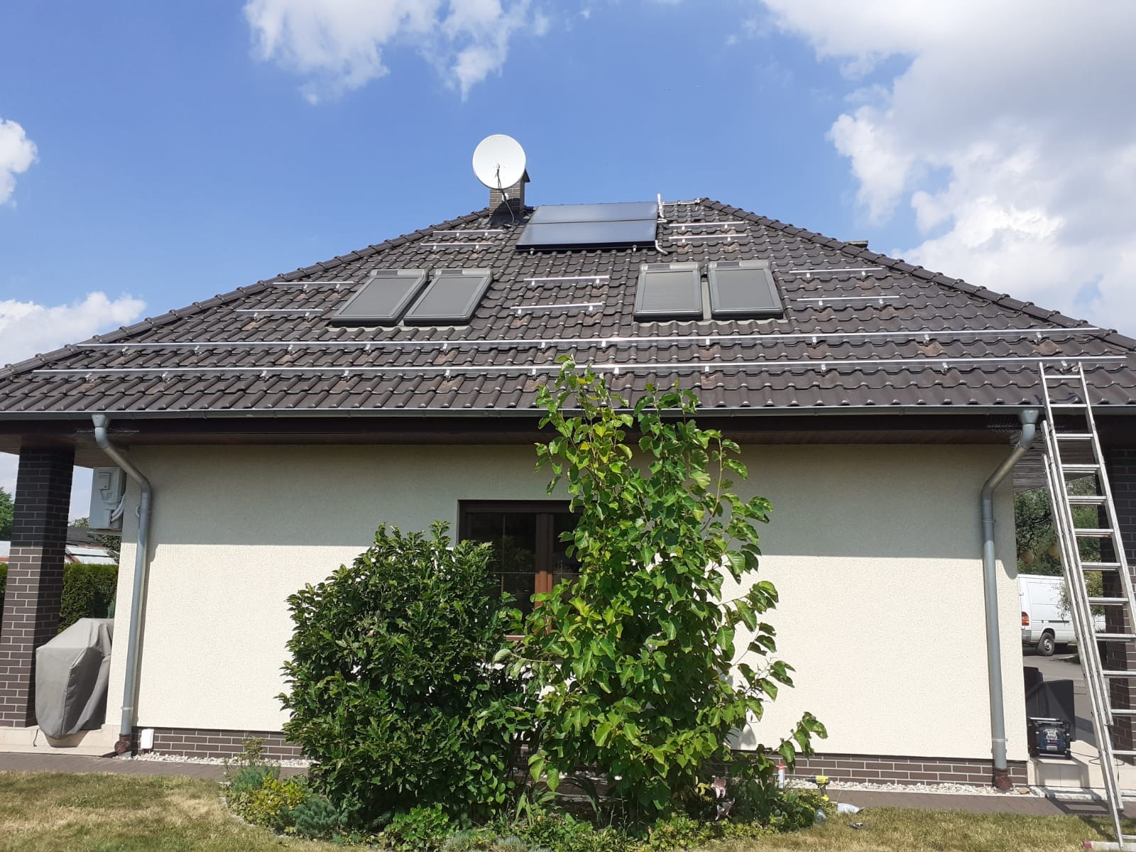 Dom jednorodzinny z zamontowanymi panelami słonecznymi na brązowym dachu, widoczna antena satelitarna i drabina oparta o rynnę.