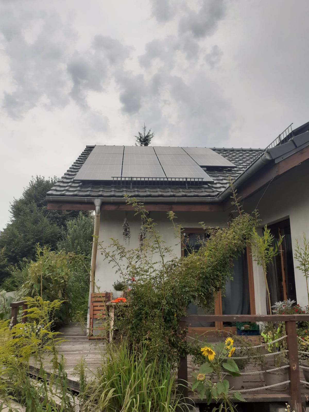 Panel słoneczny na dachu domu z drewnianym tarasem, widok z dołu na tle pochmurnego nieba, zarośnięty ogród wokół tarasu.