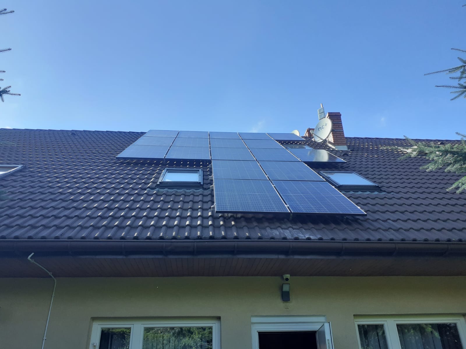 Panele fotowoltaiczne zamontowane na brązowym dachu domu jednorodzinnego z dwoma oknami dachowymi, widok z dołu w słoneczny dzień.