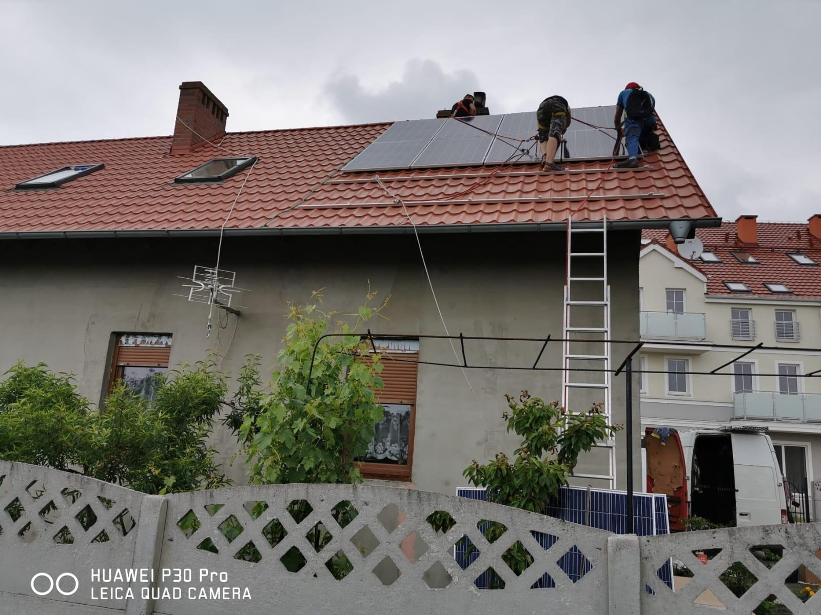 Montaż paneli słonecznych na dachu domu z czerwonej dachówki, widoczni trzej monterzy przy pracy, drabina oparta o ścianę, na pierwszym planie betonowy płot z roślinnością.