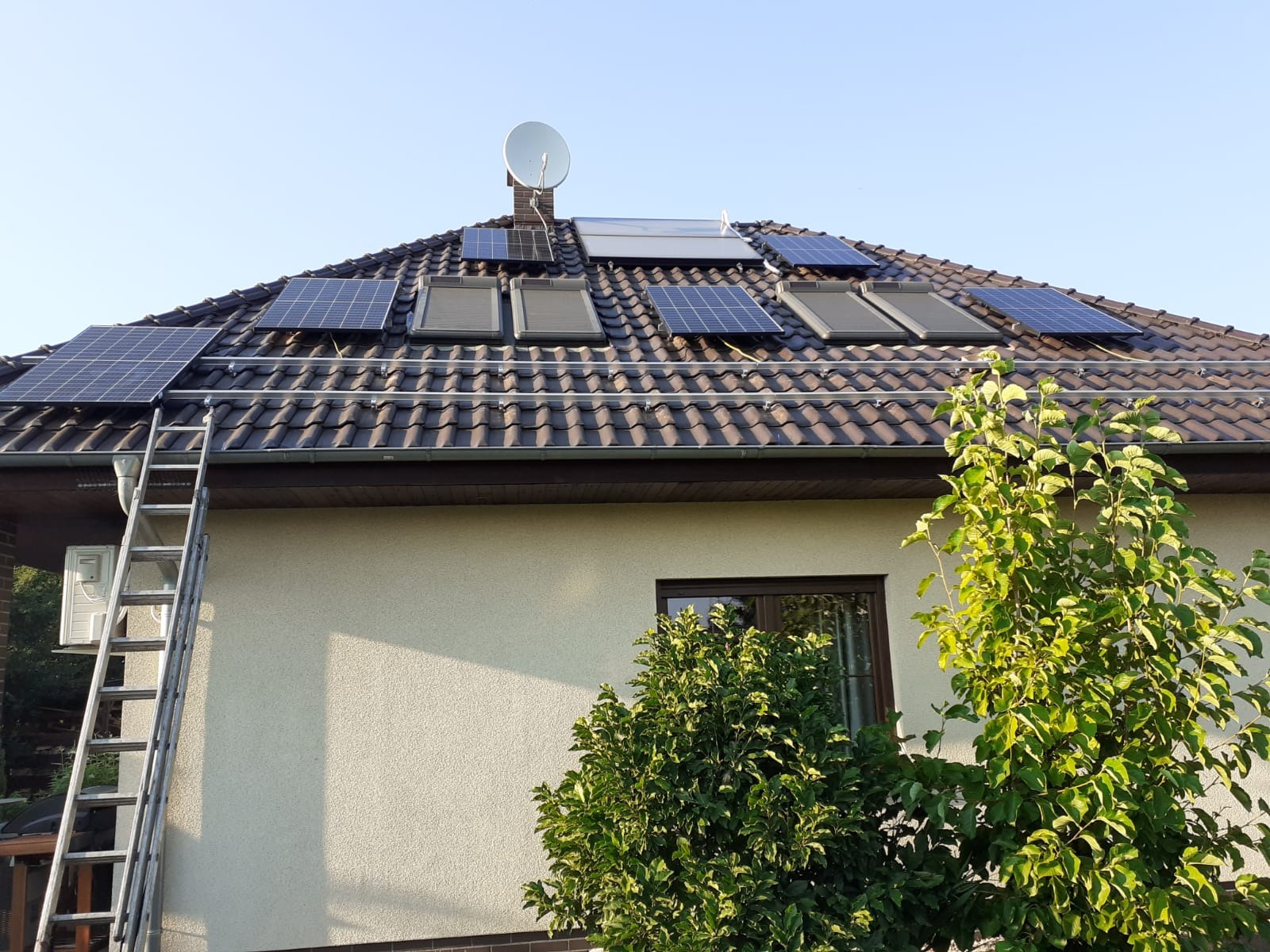 Panele słoneczne i kolektory słoneczne zamontowane na brązowym dachu domu jednorodzinnego w Polanicy-Zdroju, z widoczną drabiną opartą o ścianę i anteną satelitarną na kominie.
