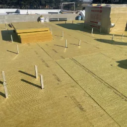 Płaski dach pokryty warstwą izolacji z wełny mineralnej, z wystającymi pionowymi wspornikami, w tle widoczne paczki wełny Rockwool i drzewa.