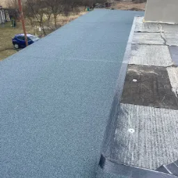 Nowe, szare pokrycie dachowe z posypką mineralną na dużej powierzchni dachu, widoczne fragmenty starego pokrycia przy kominie, w tle zabudowania i drzewa.