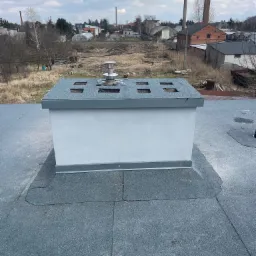 Szary dach pokryty papą z kominem wentylacyjnym na tle zabudowań przemysłowych i pochmurnego nieba.