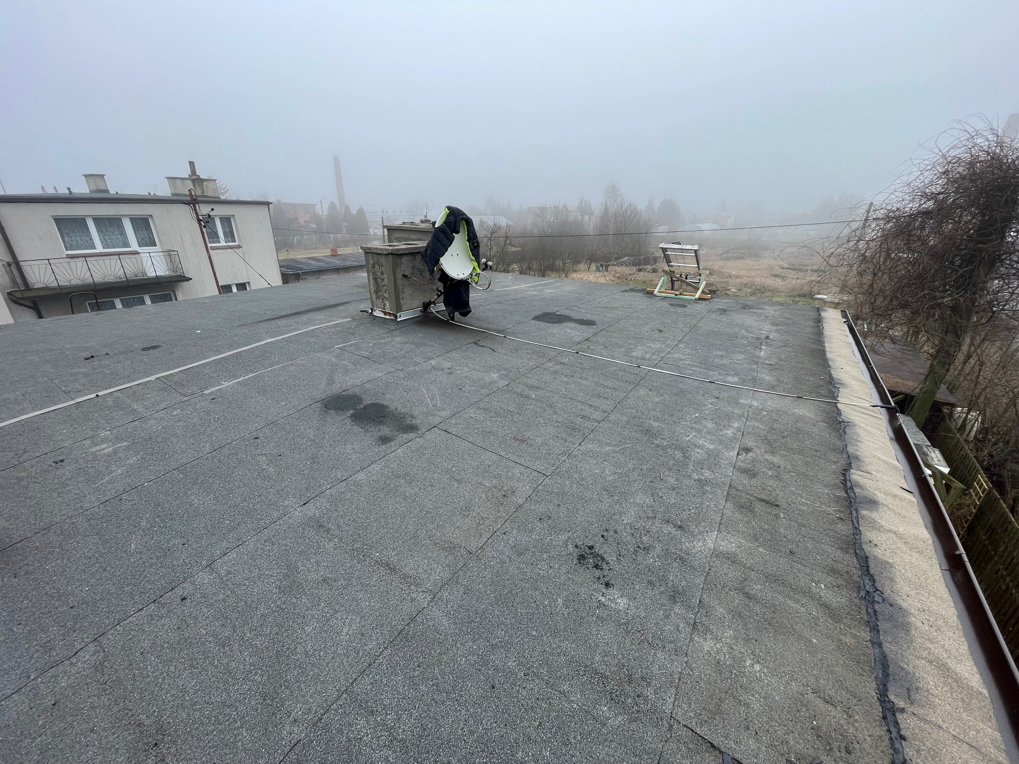 Pracownik w czarno-żółtej kamizelce na płaskim dachu pokrytym papą, obok komina wentylacyjnego i drabiny, w tle zamglony krajobraz miejski.