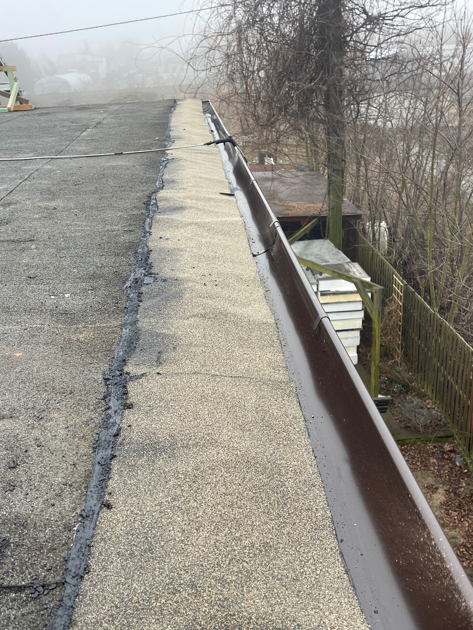 Płaski dach pokryty papą z widoczną linią łączenia i brązową rynną w pochmurny dzień.