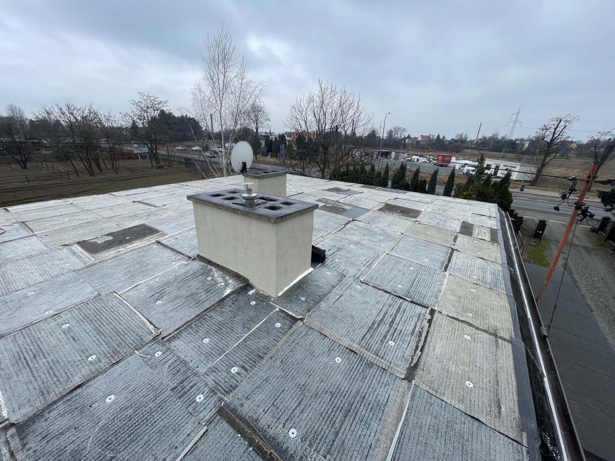 Dach pokryty papą z widocznymi łącznikami, dwa kominy betonowe z nasadami, tło z drzewami bezlistnymi i szarą zabudową miejską w pochmurny dzień.