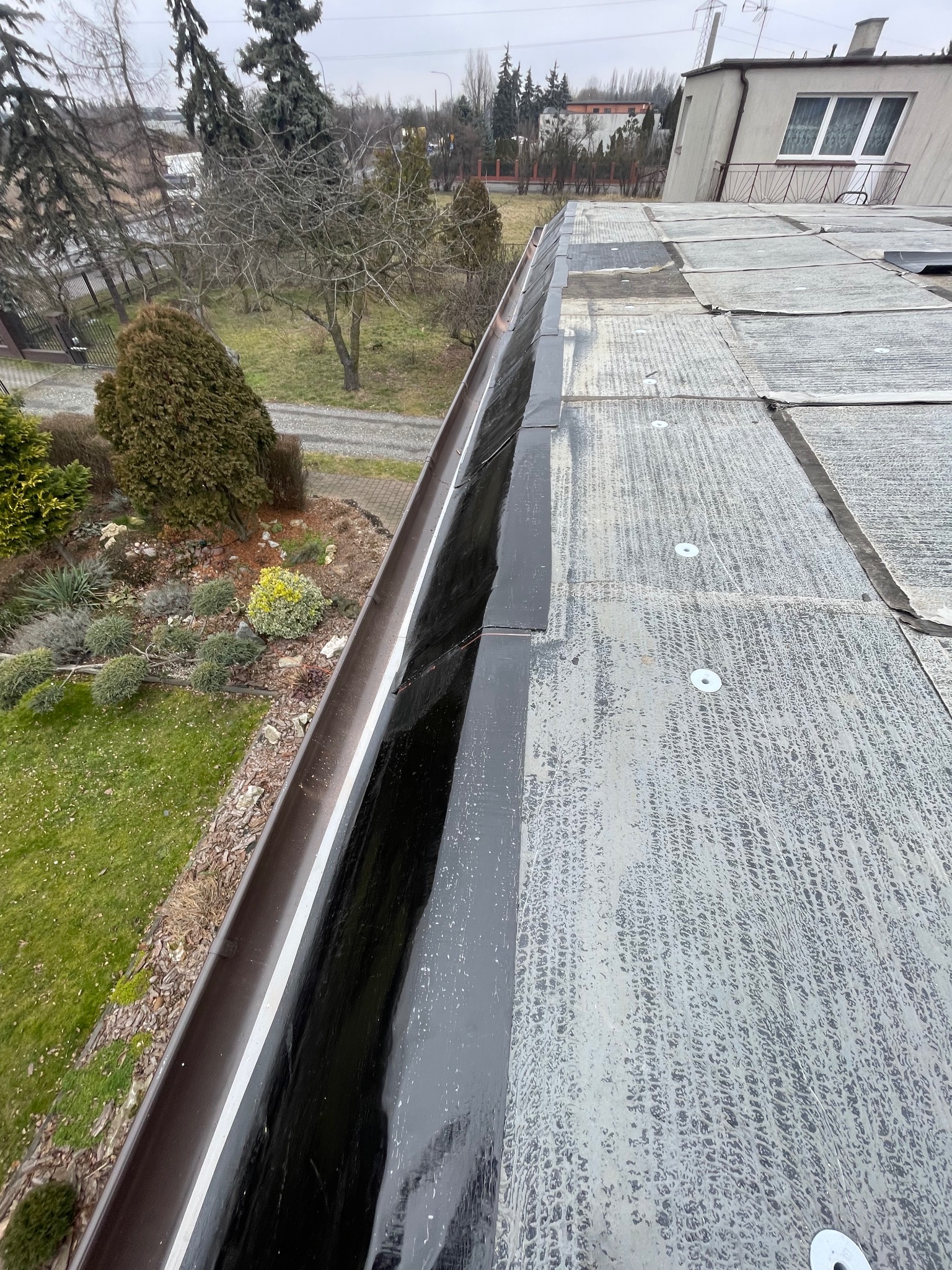 Widok z góry na płaski dach pokryty papą z widocznymi łatami i elementami mocującymi, z brązową rynną wzdłuż krawędzi i zielonym ogrodem w tle.