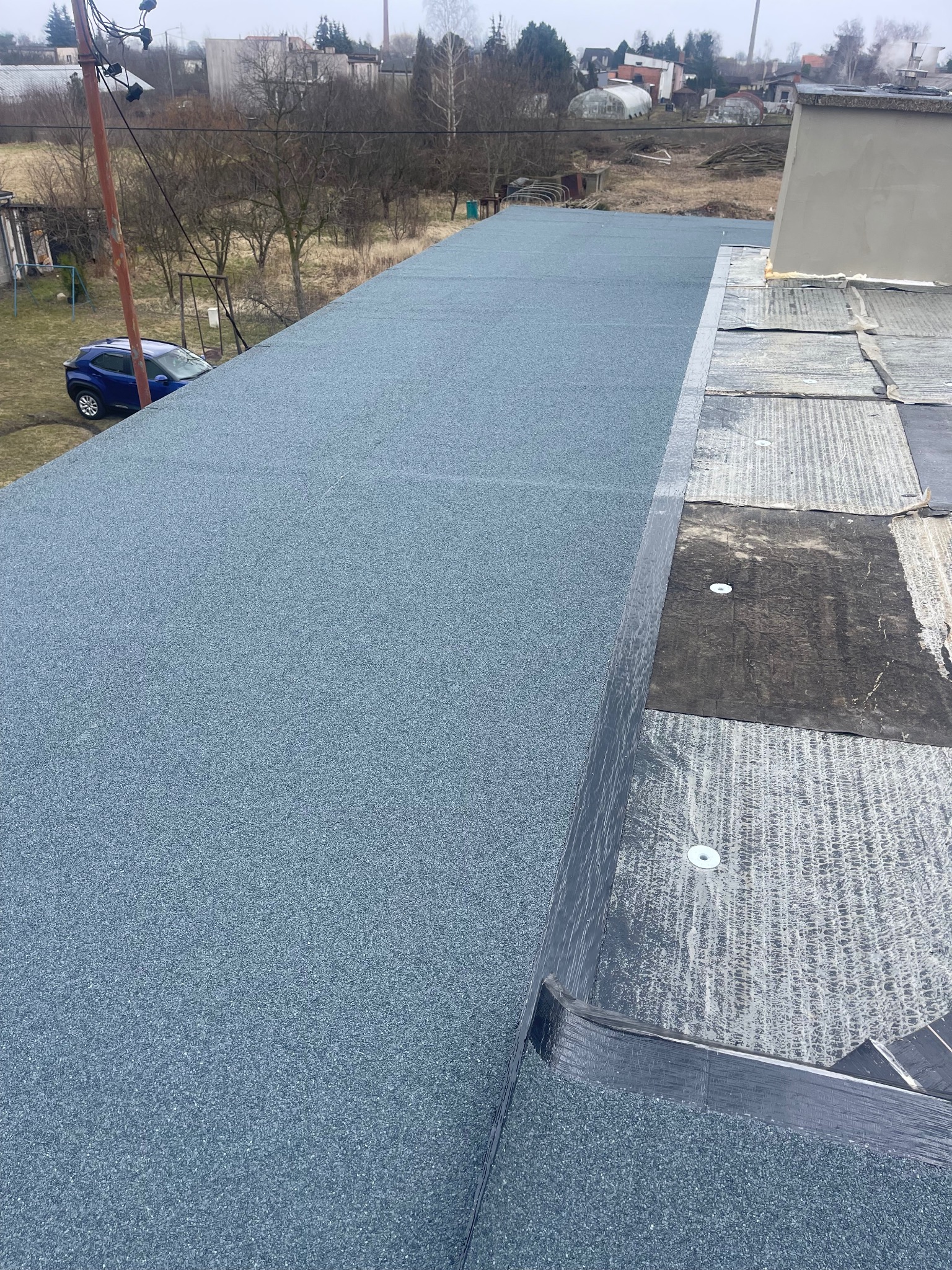 Nowe, szare pokrycie dachowe z posypką mineralną na dużej powierzchni dachu, widoczne fragmenty starego pokrycia przy kominie, w tle zabudowania i drzewa.