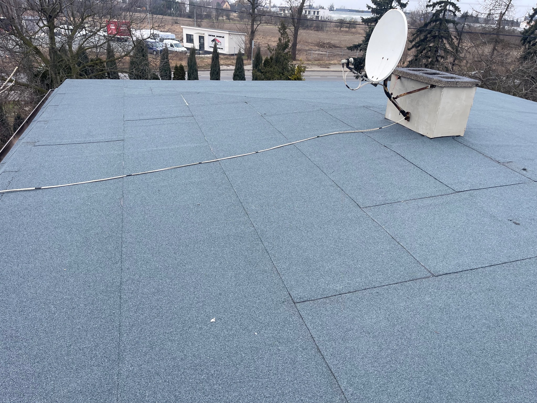 Szary dach pokryty papą termozgrzewalną, widoczny komin z anteną satelitarną, tło z drzewami i budynkami.