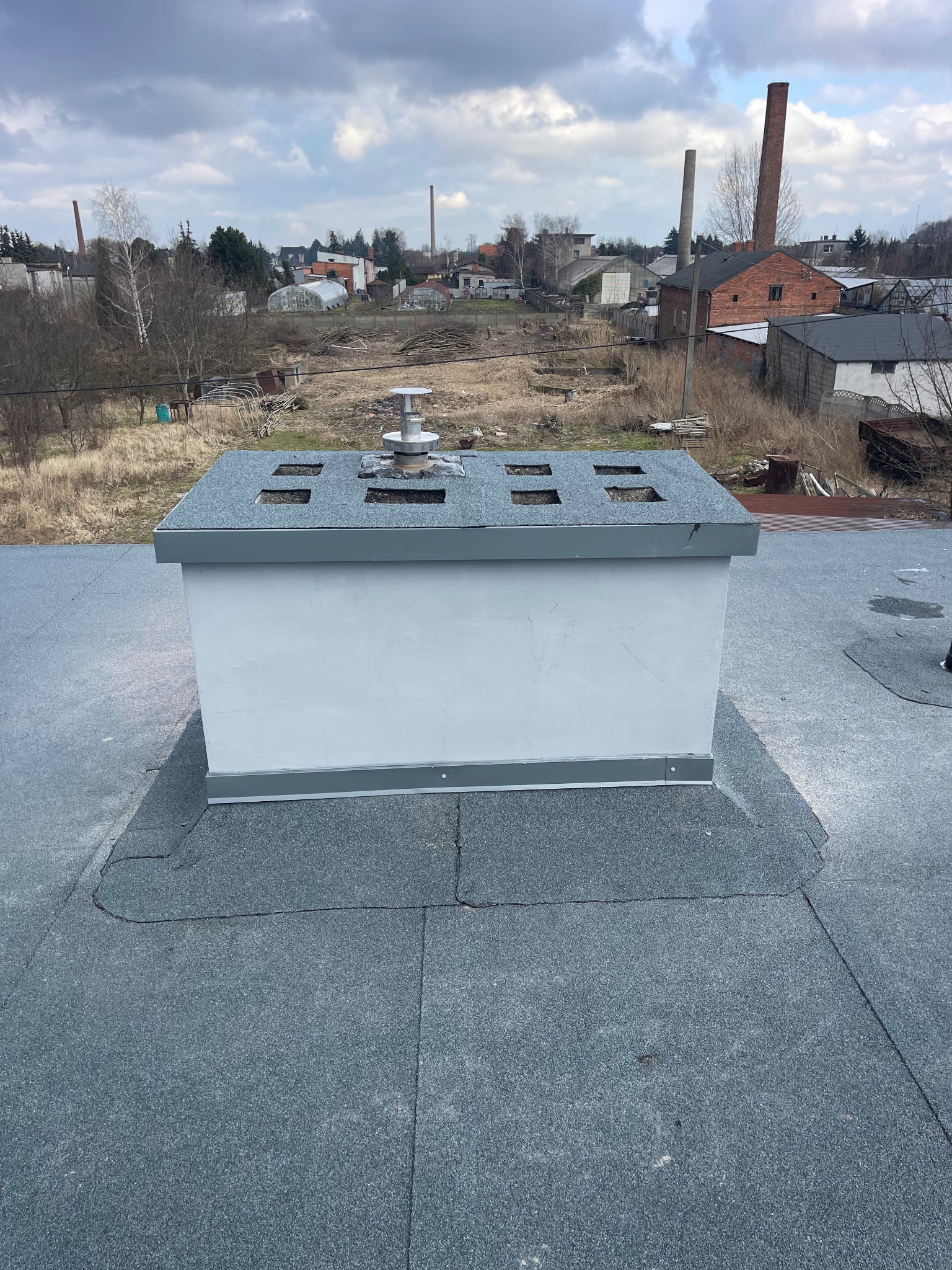 Szary dach pokryty papą z kominem wentylacyjnym na tle zabudowań przemysłowych i pochmurnego nieba.