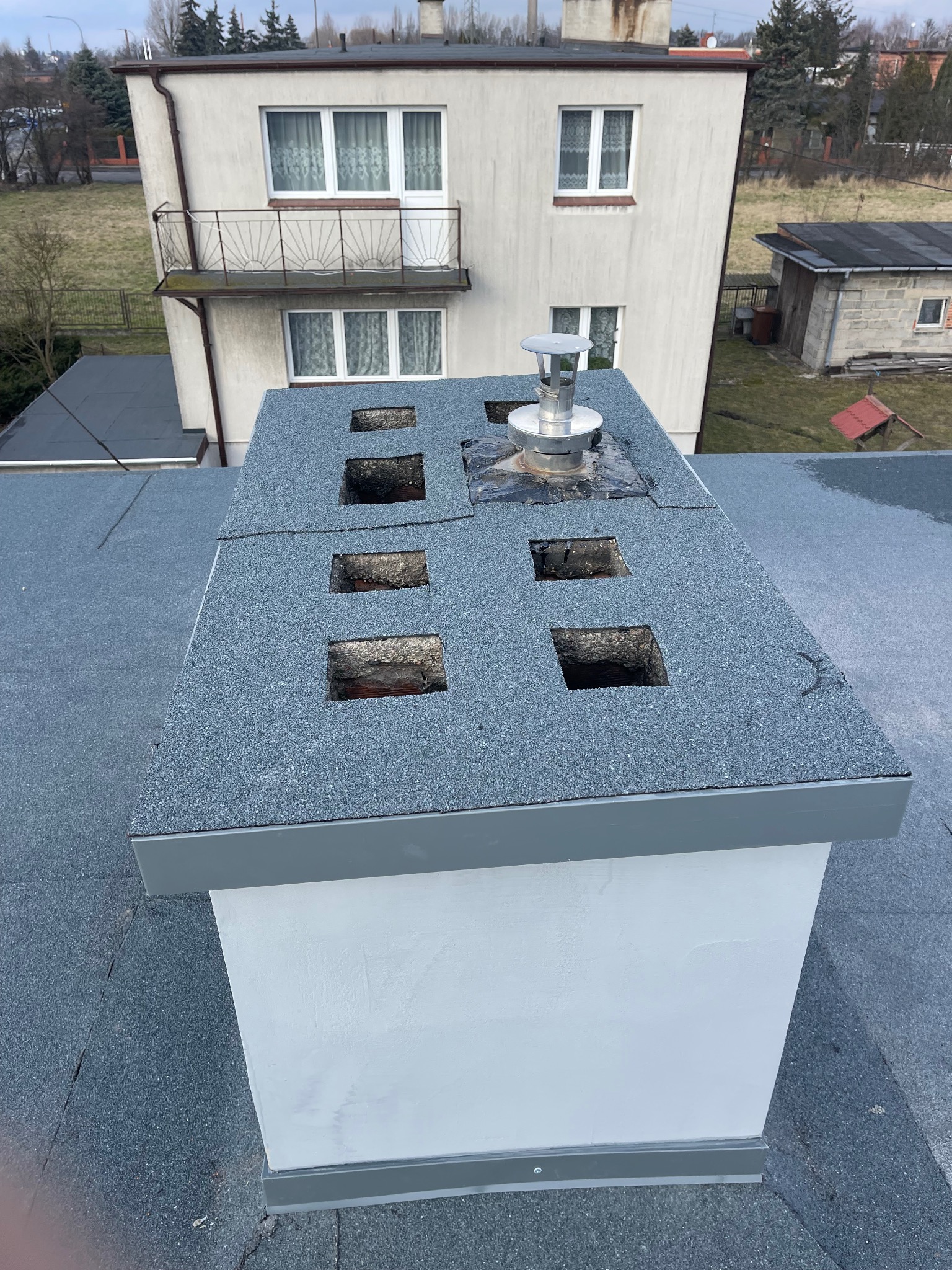 Płaski dach pokryty papą z wyciętymi otworami wokół komina, na którym zamontowany jest metalowy wiatrołap, widok z góry na budynek mieszkalny w tle.