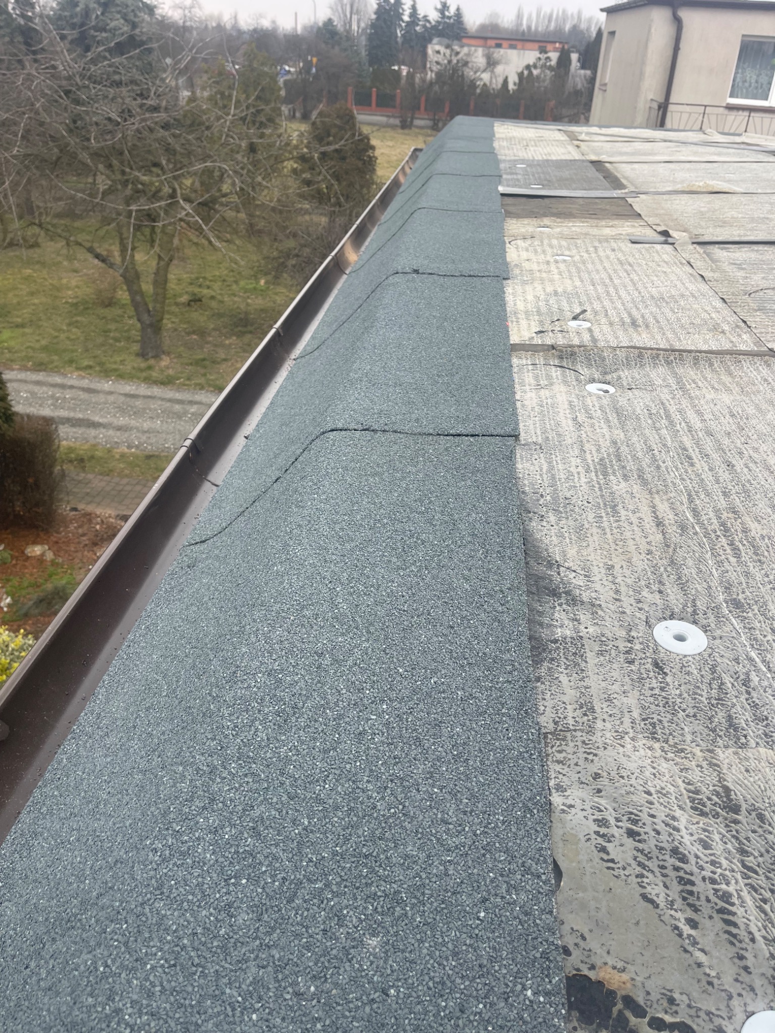 Fragment dachu płaskiego w trakcie prac: nowa papa bitumiczna z posypką mineralną sąsiaduje ze starą, przygotowaną powierzchnią, widoczne mocowania mechaniczne. Brązowa rynna wzdłuż krawędzi.