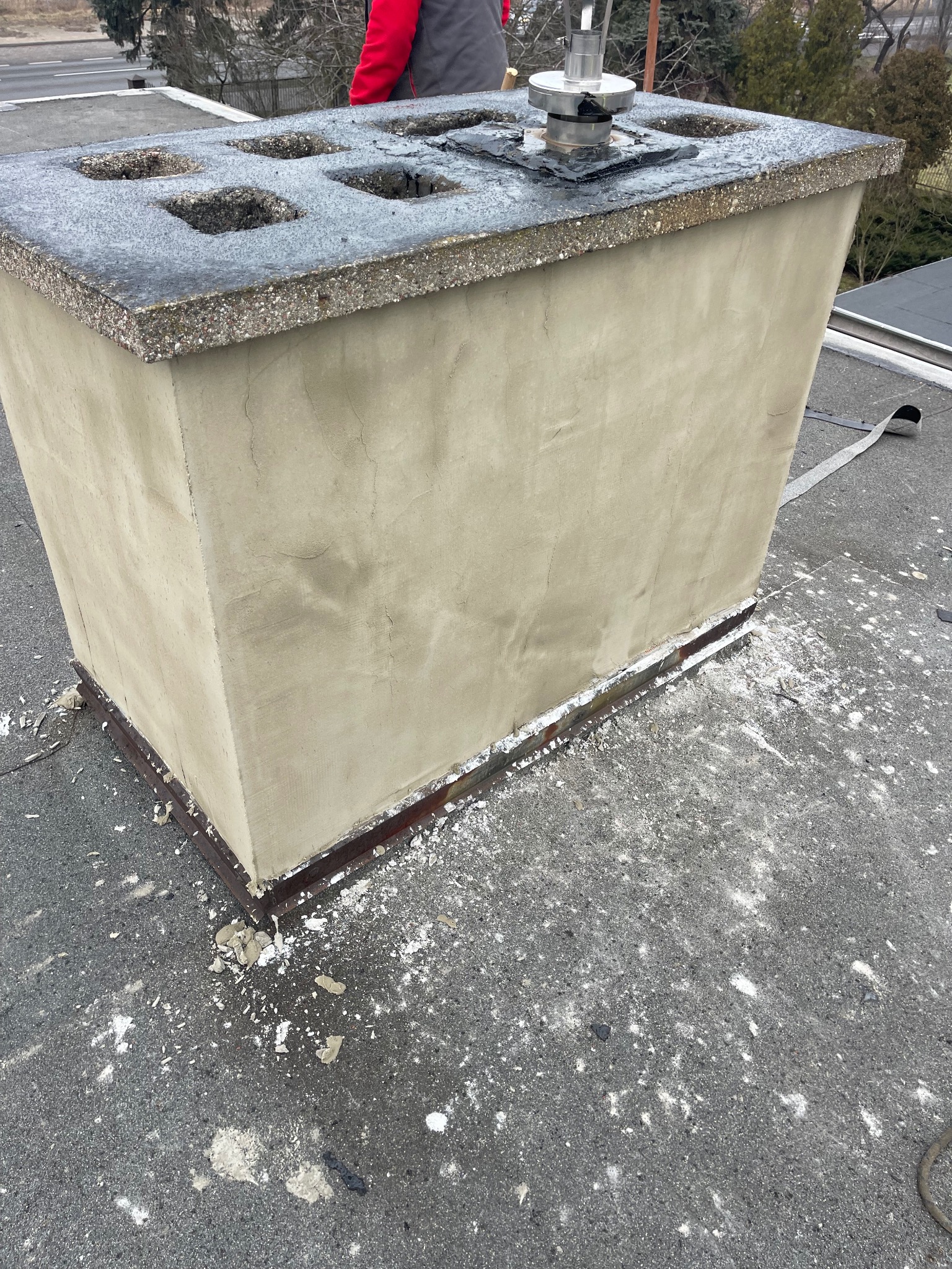 Betonowy komin wentylacyjny na płaskim dachu z widocznymi śladami napraw, fragmenty zaprawy wokół podstawy, elementy instalacji wentylacyjnej na górze.