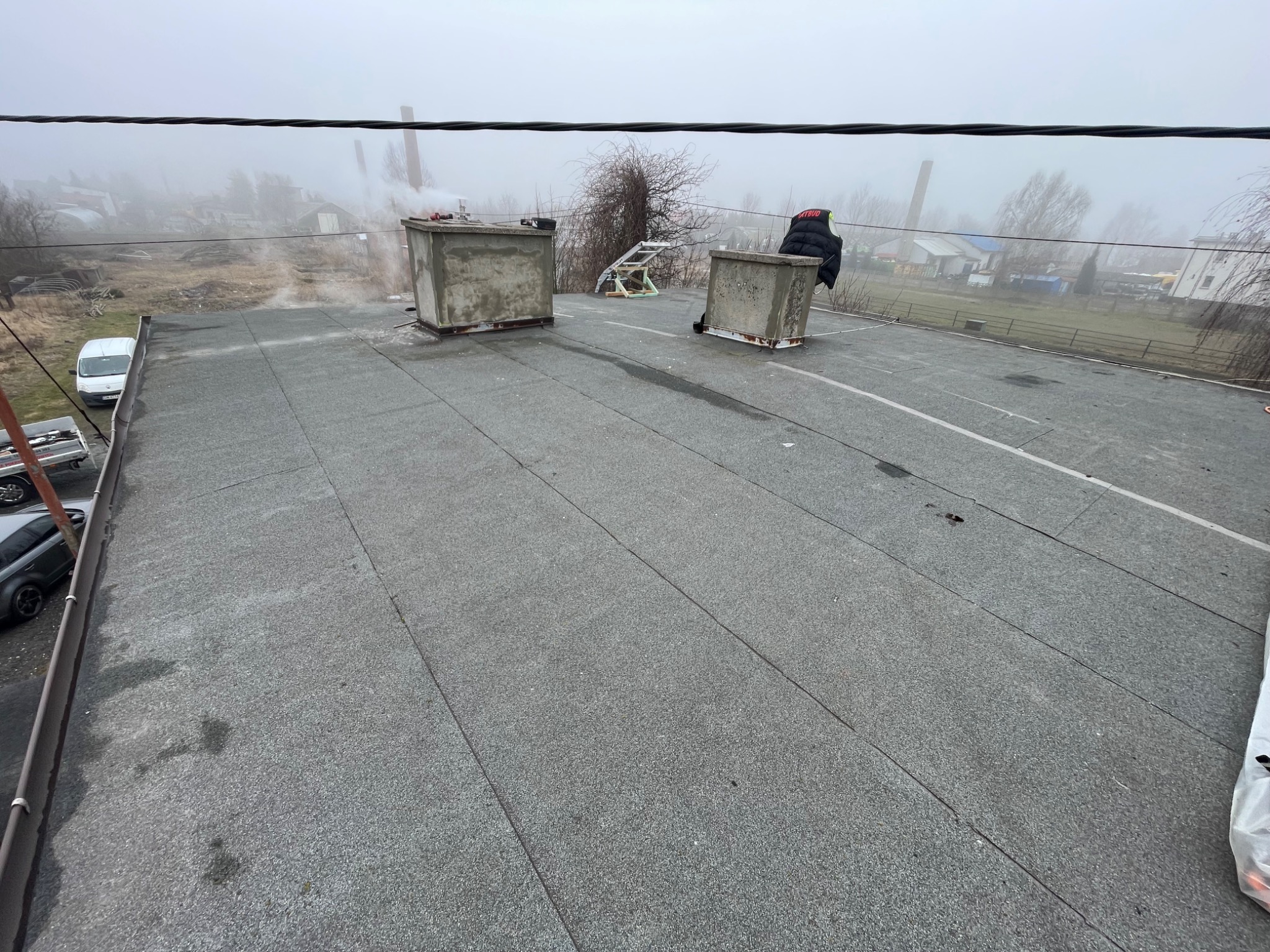 Widok z góry na płaski dach pokryty papą termozgrzewalną, z dwoma kominami wentylacyjnymi, drabinką i odłożoną kurtką roboczą w tle zamglony krajobraz.
