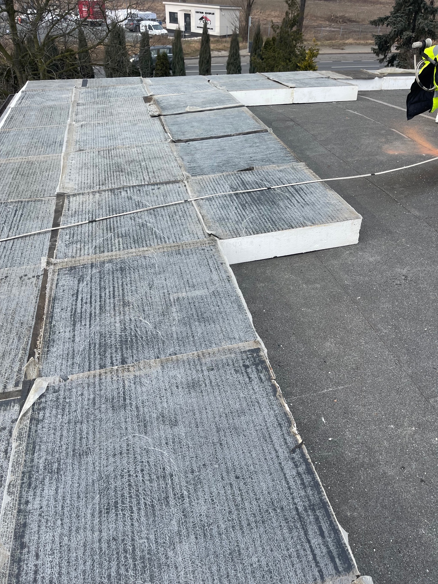 Płaski dach pokryty płytami izolacyjnymi z widocznym pracownikiem w odblaskowej kamizelce, przygotowującym się do dalszych prac.