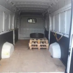 Pusta przestrzeń ładunkowa białego furgonetki z paletą i oponą w środku, widok od tyłu przez otwarte drzwi.