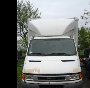 Biały samochód dostawczy marki Iveco z zabudową kontenerową, widoczny od przodu, z lekkimi śladami użytkowania na karoserii i zielonymi drzewami w tle.