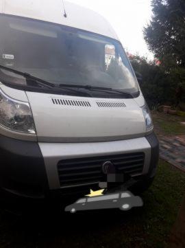 Biały bus dostawczy marki Fiat Ducato, zaparkowany na trawniku, z zamazaną tablicą rejestracyjną, widok z przodu, lekko od dołu.
