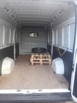 Pusta przestrzeń ładunkowa białego furgonetki z paletą i oponą w środku, widok od tyłu przez otwarte drzwi.