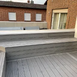 Nowoczesny taras z kompozytowych desek w odcieniach szarości, z niskimi schodami i siedziskiem, na tle ceglanego budynku z białymi roletami.