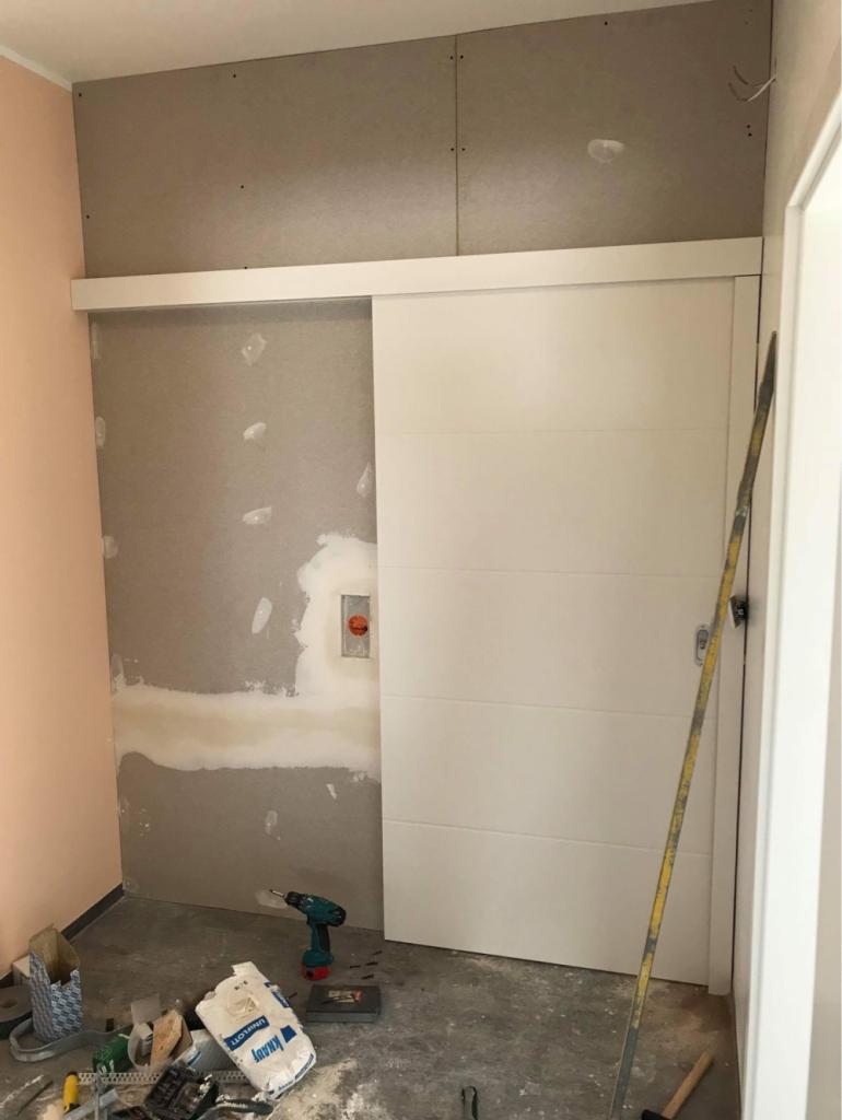 Montaż płyty kartonowo-gipsowej: widoczna ściana z płytą, częściowo pokryta masą szpachlową, białe przesuwne drzwi i narzędzia pracy, w tym wiertarka i worek ze szpachlą.