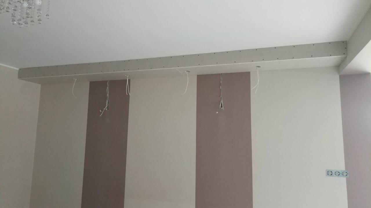 Wnętrze pokoju w trakcie prac wykończeniowych, widoczna konstrukcja z płyt karton-gips na suficie z wystającymi przewodami elektrycznymi, ściany pomalowane w pionowe pasy w odcieniach beżu i brązu,...