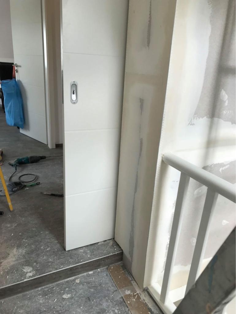 Montaż białych przesuwnych drzwi wewnętrznych z widocznymi śladami szpachlowania na ścianie i podłodze, narzędzia pracy w tle.