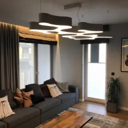 Nowoczesny salon z szarą sofą, drewnianym stolikiem kawowym na kółkach i designerskim oświetleniem sufitowym w kształcie chmur.