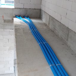 IK-METAL PROSTA SPÓŁKA AKCYJNA - Niebieskie rury instalacyjne, przymocowane metalowymi obejmami, ułożone w surowym wnętrzu budynku z białych bloczków. Widoczne otwory w ścianie.