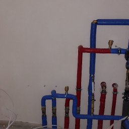 IK-METAL PROSTA SPÓŁKA AKCYJNA - Instalacja grzewcza: rury w izolacji (czerwone i niebieskie) prowadzone wzdłuż ściany, z zaworami i pompą obiegową, obok biały zasobnik ciepłej wody.