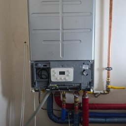 IK-METAL PROSTA SPÓŁKA AKCYJNA - Nowoczesny piec gazowy dwufunkcyjny, z widocznymi rurami instalacji CO i CWU, zaworem gazowym z żółtą rączką i fragmentem ściany.