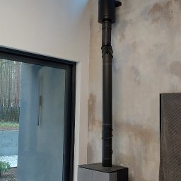 IK-METAL PROSTA SPÓŁKA AKCYJNA - Nowoczesny piec wolnostojący z czarnym kominem w minimalistycznym wnętrzu. Widoczne płomienie i drewno obok pieca. Surowy beton na ścianie dodaje industrialnego charakteru.