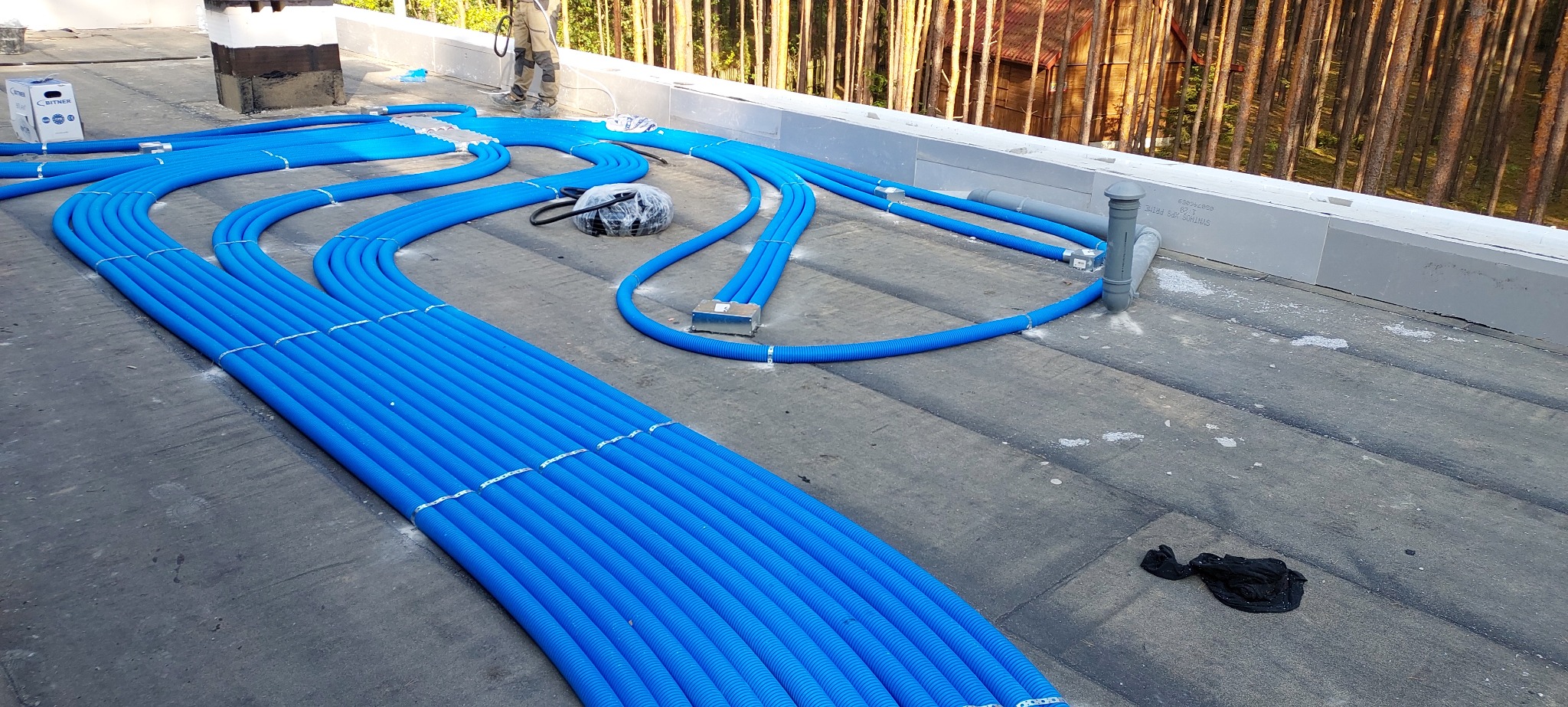 Niebieskie rury instalacji grzewczej ułożone równolegle na dachu budynku, widoczne elementy montażowe i fragment lasu w tle. Instalacja w trakcie realizacji.