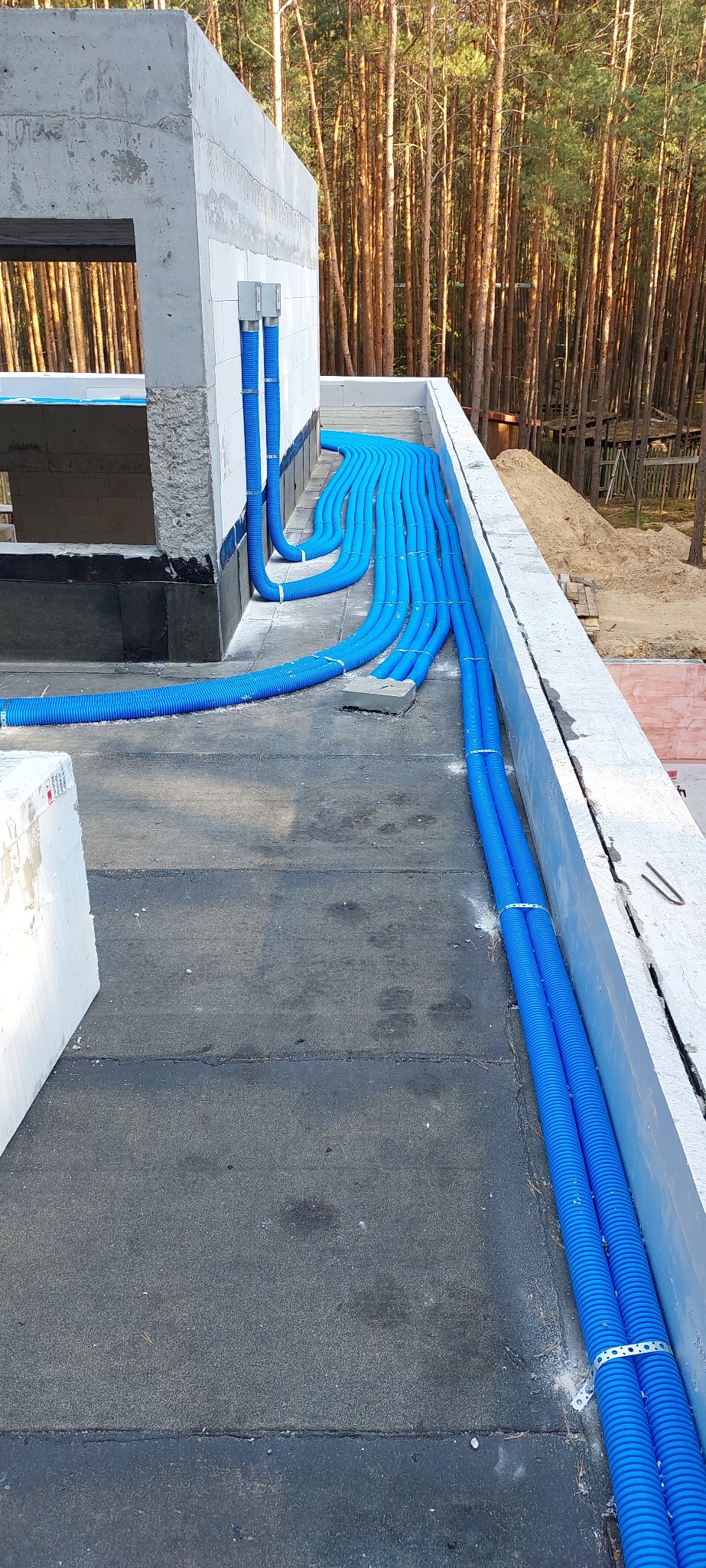 Niebieskie rury wentylacyjne ułożone na dachu budynku w trakcie budowy, widoczne mocowania i fragment ściany z otworami. W tle las iglasty.